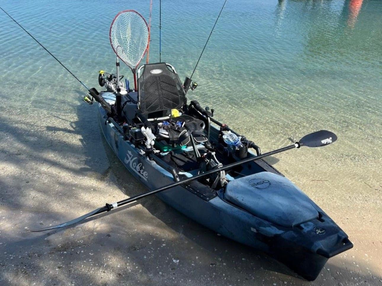 Youssef's pedal fishing kayak, rigged with rods and a landing net, pulled up on a sandy shore