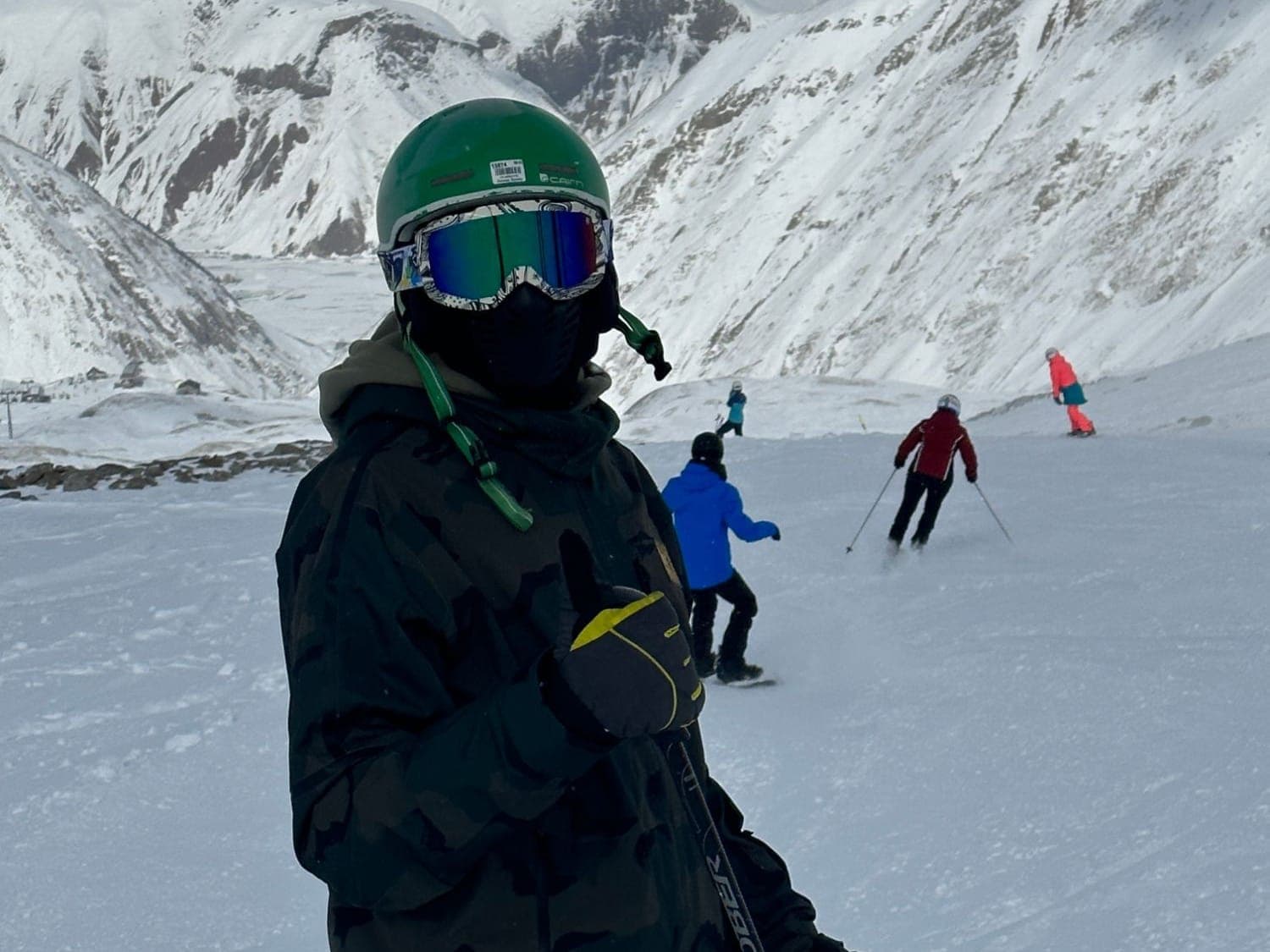 Youssef in a green helmet and goggles giving a thumbs-up on a ski slope, snowy mountains behind